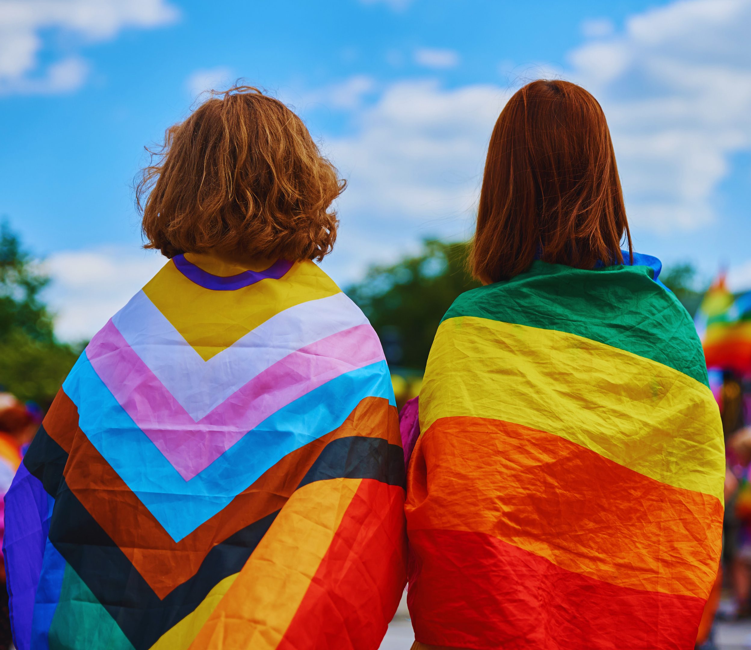 PrideFlag_GenderEqualityCause_Header Two people wear pride flags on their backs outside on a sunny day.