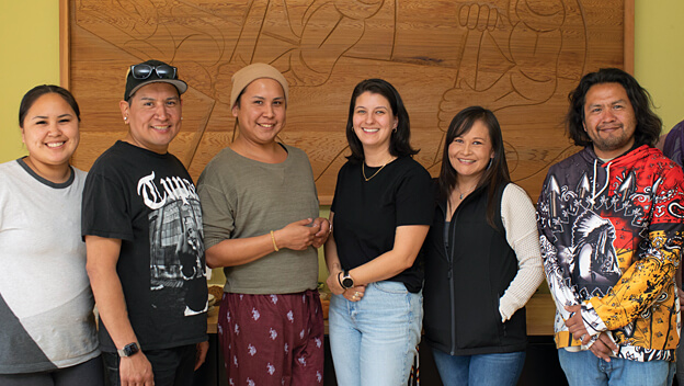 A group of people are smiling at the camera and standing in front of a large wooden Indigenous artwork.