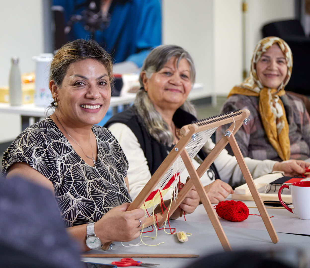 Three women look at the camera while participating in a weaving workshop at the Inter-cultural Association of Greater Victoria.