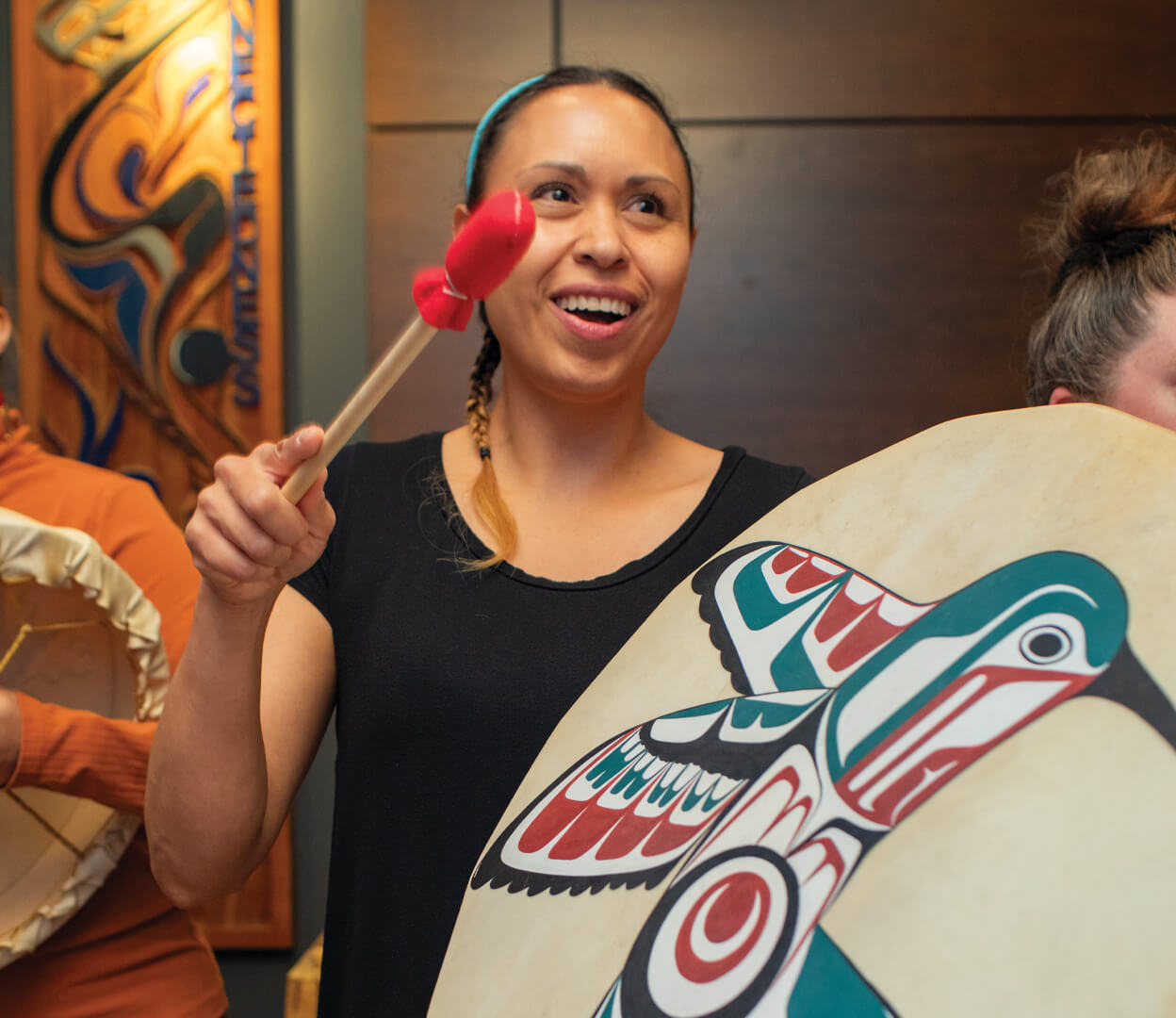 A woman is playing an Indigenous drum with a hummingbird design on it.