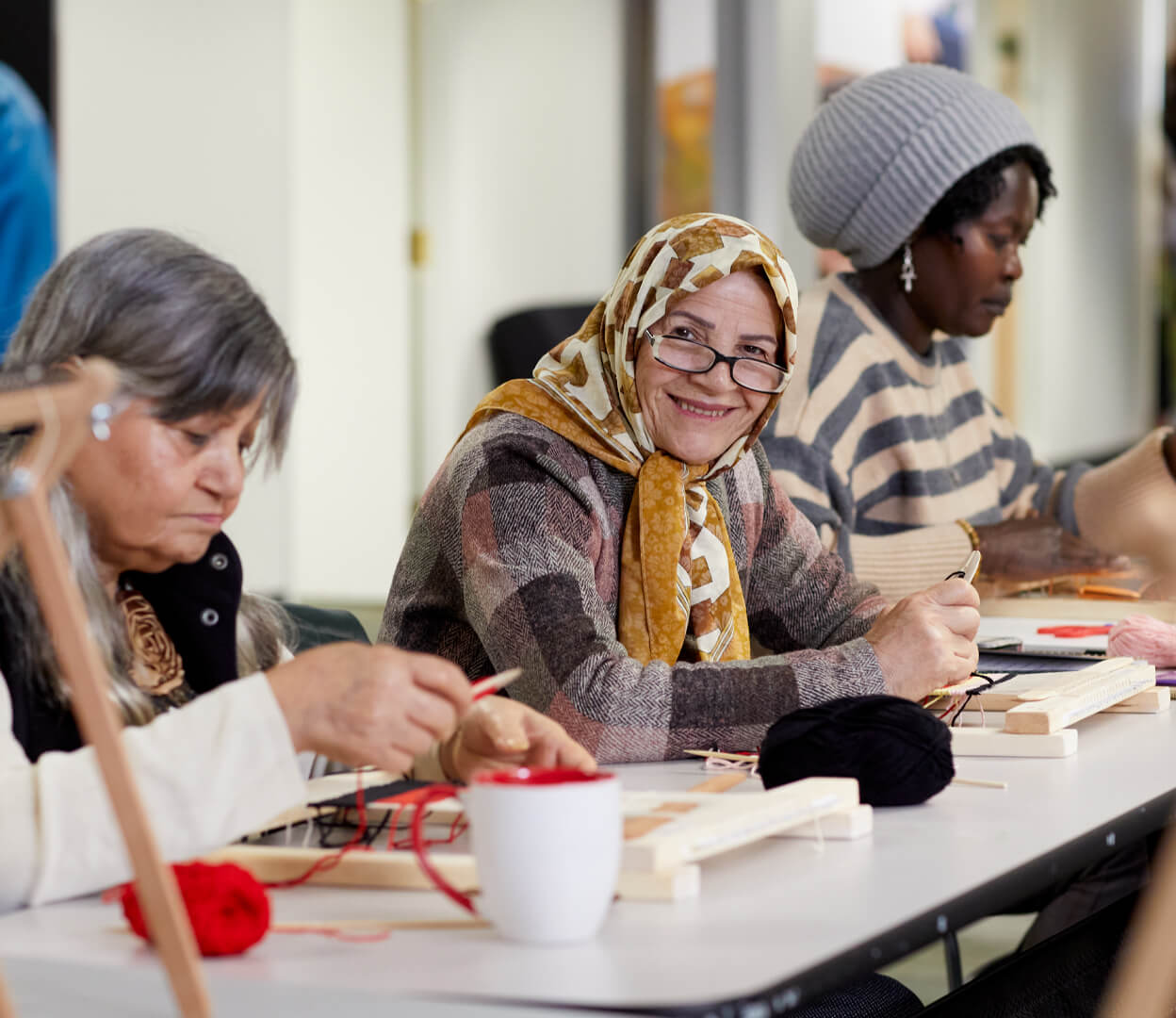 An elderly woman smiles at the camera while attending a weaving class at the Inter-cultural Association of Greater Victoria
